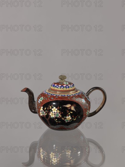 Teapot kyûsu with lid, butterflies, Japanese art, plant motifs, Japanese culture, birds, plum trees, Meiji 1868–1912, lid topped with a stylised lotus flower, cloisonné enamel. Decoration: two reserves on the belly — a plum tree and a bird; plants and butterflies; the rest of the background and the cover — a floral tendril. Date: ca. 1906–1908 Meiji 39–41. Place of origin: Kyoto, Japan. Vessel. Height 10.0 cm; diameter 10.0 cm., A rounded teapot with a short curved spout and a looped handle sits against a plain gray background and casts a soft reflection on the surface below; the teapot features a predominantly dark black glossy central area surrounded by reddish-brown enamel decorated with floral motifs in pale pink, white, and green, and small accents of gold and blue on the lid and upper band; a small ornate finial tops the lid in a muted gold tone, and the spout and handle show patterned dark brown detailing.