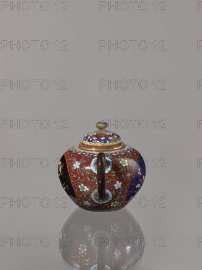 Teapot kyûsu with lid, butterflies, plant motifs, Japanese culture, birds, plum trees, Meiji 1868–1912, lids, cloisonné enamel. The lid is topped with a stylised lotus flower. Decoration: two reserves on the belly — a plum tree and a bird; plants and butterflies; the rest of the background and the cover — a floral tendril. Date ca. 1906–1908 Meiji 39–41. Japan, Kyoto. Vessel. Height 10.0 cm, diameter 10.0 cm., A small rounded lidded vessel with a central vertical handle sits on a reflective surface, showing a mirrored reflection beneath it. The vessel is richly decorated with floral and scrolling patterns in colors including red, blue, turquoise, green, pink, black, gold, and purple. The lid has concentric decorative bands and a small ornate gold-colored finial. The body displays panels of different background colors with flower motifs and curving vine designs, and the handle has a dark textured appearance. The background is a plain neutral gray.