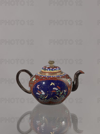 Teapot kyûsu with lid;, Cloisonné enamel; ca. 1906–1908 Meiji 39–41, Kyoto, Japan. Height 10.0 cm, diameter 10.0 cm. The lid is topped with a stylised lotus flower. Decoration: two reserves on the belly showing a plum tree and a bird, with plants and butterflies; the remainder of the background and the cover decorated with a floral tendril. Subjects: birds, plum trees, plant motifs, butterflies., A rounded teapot with a curved handle and spout sits against a gray background, showing a reflective surface beneath. The teapot features a dominant deep blue central band decorated with floral motifs in green, white, pink, yellow, and red, surrounded by reddish-brown borders with floral and leaf patterns. The lid has concentric decorative rings in blue, red, and gold topped by a small gold-colored finial. The handle and spout are dark brown with a subtle dotted texture. The overall colors visible include blue, red, brown, gold, green, white, pink, yellow, and gray.
