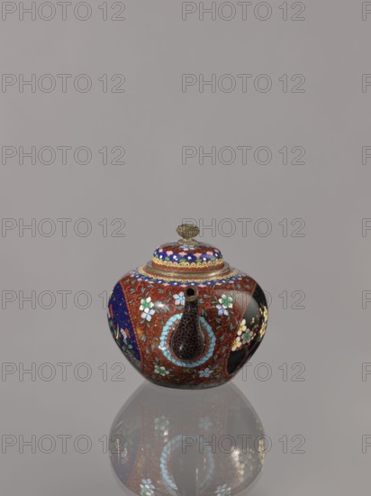 Teapot kyûsu with lid, ; cloisonné enamel, Meiji 1868–1912, ca. 1906–1908 Meiji 39–41; Japan, Kyoto. Vessel height 10.0 cm, diameter 10.0 cm. Decoration: lid topped with a stylised lotus flower; two reserves on the belly depicting a plum tree and a bird, with plants and butterflies; remaining background and cover with a floral tendril. Subjects: butterflies, birds, plum trees, plant motifs; Japanese., A small rounded decorative vessel with a lid sits centered against a plain light gray background and casts a reflection below; the vessel has a short spout and a knobbed lid and is richly patterned with floral motifs and curved designs in colors including red, blue, black, white, green, gold, and turquoise, with metallic gold accents and a dark textured area around the spout, and a glossy finish that reflects light.