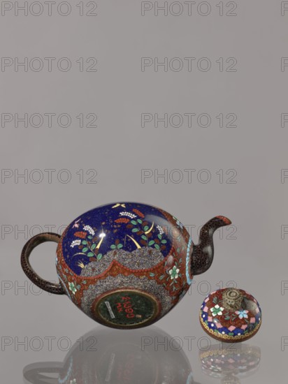 Teapot kyûsu with lid;, Cloisonné enamel teapot, ca. 1906–1908 Meiji 39–41, Kyoto, Japan. Height 10.0 cm, diameter 10.0 cm. The lid is topped with a stylised lotus flower. Decoration: two reserves on the belly—a plum tree and a bird—plus plants and butterflies; the rest of the background and the cover decorated with a floral tendril. Subjects: birds, plum trees, butterflies, plant motifs., A rounded teapot and a separate lid rest on a reflective surface against a plain grey background. The teapot has a deep blue upper surface with floral motifs in green, white, yellow, and red, and a band of textured, pebble-like patterning in grey and brown around its midsection accented with red and turquoise floral decorations. The handle and spout are dark brown with a subtle scale-like or dotted texture, and the spout tip shows a small area of red. The separate lid echoes the teapot’s colors with blue, red, turquoise, white, and gold decorative patterns and a central metallic, sculpted finial in a...