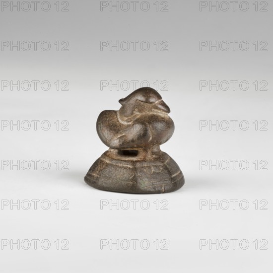 Weight in the shape of a duck standing on two short legs and a similar support, on a wide, octagonal base that widens towards the bottom. The bird is short, stocky. The head is recessed, nestled into the body, with a pointed ruff slightly upturned. The eyes are oblong and convex. The beak is broad and flat. The breast is broad and convex, and the folded wings are marked with a contour. The tail is folded, triangular, and slightly upturned. The base is decorated with continuous lines: one in the upper and two in the lower part of the side walls. A symbol is engraved on one of the base walls. Flat bottom, gently contoured edges. 17th/18th century, Burma Myanmar. Weight; height 3.8 cm, width 3.8 cm, depth 3.6 cm, weight 0.14 kg., classification: weight; motifs: birds, pharmacies;, scales., A small sculptural object sits centered on a pale surface against a light gray background; it appears carved with rounded, organic forms resembling a stylized bird or animal perched on a contoured base....