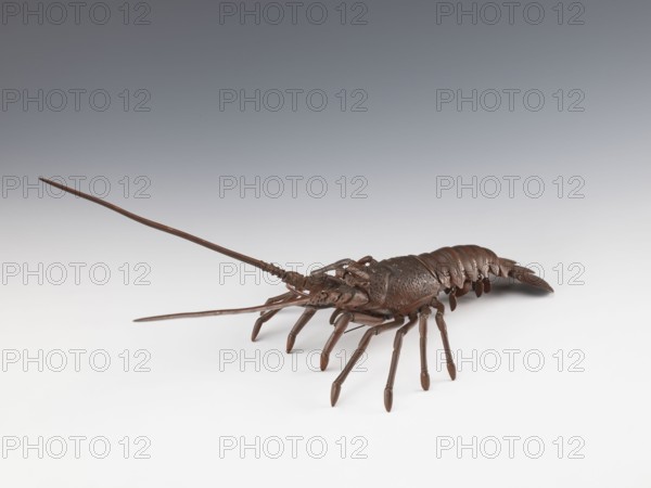 Lobster-shaped okimono Ise ebi, late 19th century Meiji period, 1868–1912, Japan. Bronze sculpture jizai okimono, depicting a lobster; length 42.0 cm, width 21.0 cm, weight 1.08 kg. This type of lobster is called Ise ebi because many are harvested in Ise Province Mie Prefecture. The lobster's spiny shell is said to protect people from demons oni and bring good luck. In some regions of Ise Province, there is an old custom of decorating shrine shimenawa sacred ropes with lobster shells. Japanese art; figurine, marine animal., A sculptural object resembling a crustacean sits on a smooth surface, rendered in a dark brown color with a slightly glossy finish; it has a segmented body with overlapping plates, a textured central carapace, multiple jointed legs extending downward, and long slender antennae projecting forward, all in the same brown tone against a background that transitions from light gray near the surface to darker gray higher up.