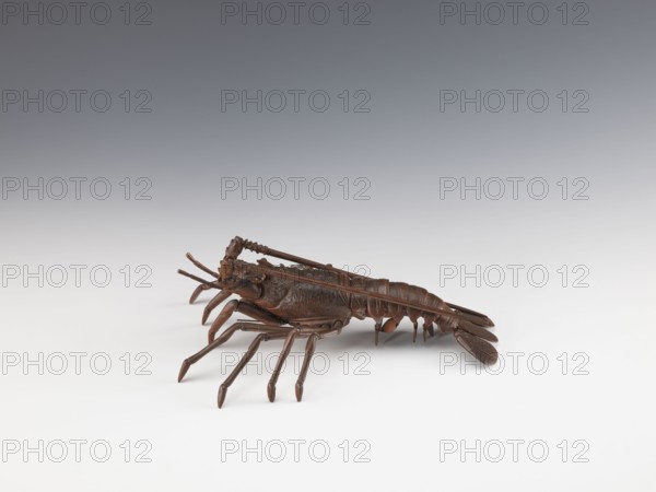 Lobster-shaped okimono Ise ebi, bronze figurine jizai okimono depicting a lobster; Meiji period, late 19th century 1868–1912, Japan. Inscription/history: This type of lobster is called Ise ebi because many are harvested in Ise Province Mie Prefecture; the spiny shell is said to protect people from demons oni and bring good luck. In some regions of Ise Province, lobster shells were used to decorate sacred ropes shimenawa at shrines. Sculpture; length 42.0 cm, width 21.0 cm, weight 1.08 kg., A sculpted brown object resembling a crustacean rests on a smooth surface with a gradient background that transitions from light gray near the surface to darker gray toward the top. The object has a segmented body, a fan-like tail, several jointed legs and antennae, and a textured surface with darker and lighter brown tones. The surface beneath and around the object is mostly white to very pale gray, and the scene is evenly lit with soft shadows beneath the object.