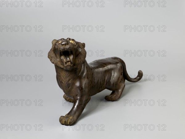 Okimono in the shape of a tiger;, Meiji period 2nd half of the 19th century, 1868–1912; Japanese; sculpture; materials not stated; figurine of an animal tiger; height 18.0 cm, width 35.0 cm, weight 3.16 kg., A sculpted animal figure resembling a roaring big cat with an open mouth, visible teeth, and a protruding tongue, shown in a three-quarter view. The figure has a muscular body, a mane-like textured area around the head, and a long curved tail. The surface shows carved fur details and smooth areas with a patinated finish. The sculpture rests on a flat surface and casts subtle shadows. The visible colors include shades of brown, dark bronze, gold highlights in the eye areas, and a light gray background.