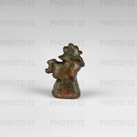 Weight; maker, purchase provenance; scales; Southeast Asia; pharmacies; lions; 17th–18th century; Burma Myanmar; weight: 0.03 kg; height: 3.0 cm; width: 2.2 cm; depth: 1.6 cm., A small sculptural object with a rough, irregular shape sits on a smooth light gray surface against a pale gray background; the object has a predominantly brown and dark brown surface with patches of green patina, an uneven silhouette with bulbous and rounded protrusions, and a slightly flared base that supports the irregular upper form.