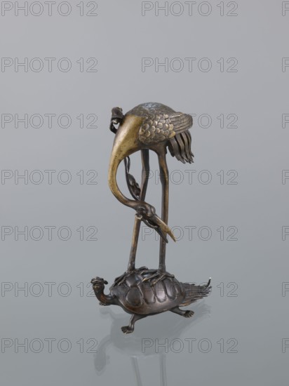 Candlestick shokudai, lotuses, turtles, cranes iconography, Japanese art, Meiji 1868–1912, animals, 2nd half of the 19th century, Japan, candlestick, height 37.0 cm, A metallic sculpture showing a tall bird standing on a smaller animal; the bird has a long curved neck and beak, textured wings with layered feather details, slender legs and clawed feet, and a patina varying between golden-bronze and darker brown tones, while the smaller animal beneath has a domed shell with segment-like ridges, a small head with a beak-like mouth, short legs, and a darker brown metallic finish; the background is a smooth light gray and the surface beneath the sculpture reflects faintly.