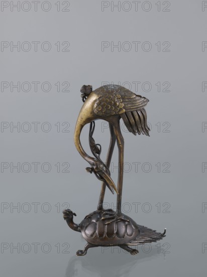 Candlestick shokudai, ; materials/technique: metal with decorative motifs of lotuses, turtles, and cranes; subject/iconography: animals and lotuses with Genius figure; style/period: Japanese, Meiji 1868–1912, 2nd half of the 19th century; function: candlestick; height: 37.0 cm., A metal sculpture showing a long-legged bird standing on the back of a turtle, both rendered with textured details; the bird has a curved neck and a long beak with layered feather patterns on its body and tail, and the turtle has a domed shell with scalloped segments and an extended head and legs. The sculpture appears in metallic tones of dark bronze and golden-brass against a neutral light gray background.
