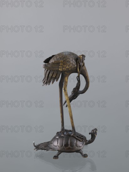 Candlestick shokudai, ; materials and iconography: lotuses, turtles, cranes, animals, depiction of a Genius; Japanese, Meiji 1868–1912, 2nd half of the 19th century; height 37.0 cm., A metallic sculpture shows a bird with long thin legs standing on the back of a turtle; the bird has textured wings with layered feathers and a curved beak, and some dangling feather-like elements hang from its body. The turtle has a domed shell with segmented plates, small legs, and a raised head with a rounded snout. The sculpture displays metallic tones including dark brown, bronze, and hints of gold against a smooth neutral gray background.