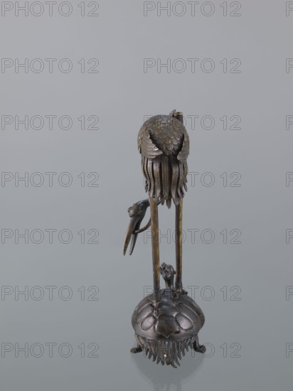 Candlestick shokudai, lotuses, turtles, cranes iconography, Japanese art, Japanese culture, Meiji 1868–1912, animals, 2nd half of the 19th century, Japan, candlestick, height 37.0 cm, A tall metallic sculpture with a dark bronze and brass patina stands on a rounded base resembling a shell with textured ridges and fringe-like elements, supported by small clawed feet; two long, slender vertical legs rise from the base to support a rounded upper form covered in overlapping scale-like plates and flowing, ribbon-like strands that hang downward, with a curved beak-like element attached to one side; the colors visible are dark bronze, brown, brassy gold, and muted black against a pale gray background.