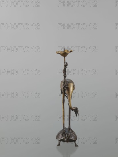 Candlestick shokudai, ; materials/features: lotuses, turtles, cranes iconography, animals; culture: Japanese, Meiji 1868–1912, 2nd half of the 19th century, Japan; height 37.0 cm., A slender metal sculpture with a muted gold and dark bronze finish stands on a rounded base that resembles a turtle form with textured shell-like details and small feet; two long, thin vertical supports rise from the base, one of which curves into a drooping elongated shape that ends in a clawed foot, while a vine-like element wraps partway up the supports and carries a small decorative flower near the top; the uppermost section features a shallow petal-shaped dish with a scalloped edge in a warm gold tone; the background is a smooth, neutral gray and the surface beneath the sculpture shows a soft reflection.