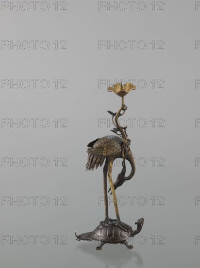 Candlestick shokudai, ; materials not specified; decorated with lotuses, turtles, cranes and depictions of a genius; Japanese, Meiji period 1868–1912, 2nd half of the 19th century; Japan; candlestick; height 37.0 cm, A metallic sculpture of a bird standing on a turtle, with the bird rendered in a gold and dark brown metal finish and the turtle in a darker brown metal; the bird has long slender legs, a curved neck, detailed feather textures on its body and a cluster of elongated tail feathers, and it supports a winding vine-like stem that rises upward to end in a flower-shaped cup with a gold tone; the turtle has a domed, segmented shell, short legs, a visible head and tail, and both figures cast faint reflections on a smooth gray surface with a plain light gray background.