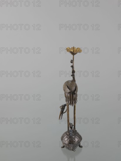 Candlestick shokudai, lotuses, turtles, cranes iconography, Japanese art, Japanese culture, Meiji 1868–1912, animals, 2nd half of the 19th century, Japan, candlestick, height 37.0 cm, A slender metallic sculpture stands against a pale gray background, showing a vertical form with a base resembling a rounded shell or dome with textured segments and fringe-like elements at its lower edge; from this base rise long thin supports leading up to a bell-shaped cluster of layered, petal-like pieces and dangling fringe, with a small bird-like figure perched on one of the supports; higher up a twisted vine-like stem with tiny leaf shapes ascends to a flared, shallow cup at the top. The visible colors are bronze, gold, dark brown, and muted gray.