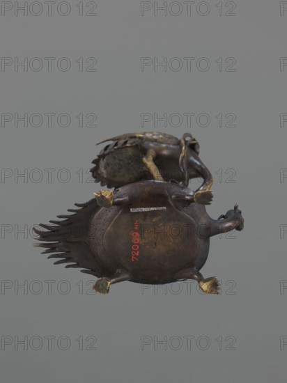 Candlestick shokudai, lotuses, turtles, cranes iconography; Japanese art, Japanese; Meiji 1868–1912, 2nd half of the 19th century, Japan; candlestick, height 37.0 cm., A sculptural object resembling an animal with a rounded dark brown body and a ridged tail is shown against a plain gray background, featuring a glossy dark bronze surface with areas of lighter brown patina and worn metallic highlights. The piece has a pair of stubby legs with gold-toned feet, a small curved head with a short snout, and decorative ridged elements along the tail that are darker brown to nearly black. A second similar object is partially visible behind the main piece, showing similar dark brown and gold tones and textured surfaces. The overall colors visible are shades of dark brown, black, gold, and gray.