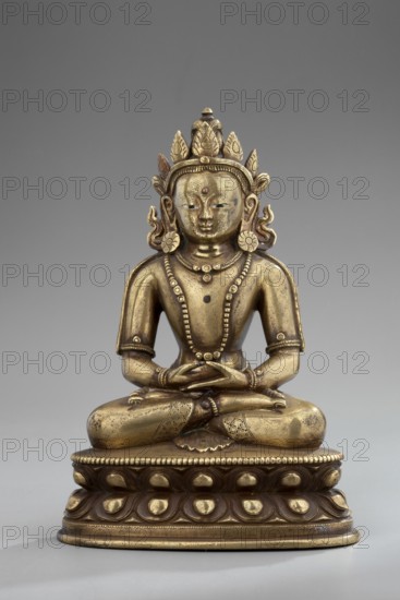 Amitayus, Himalayan/Tibetan Buddhist and Chinese/Mongolian cultural influences, lotus throne iconography; Buddha Amitayus seated on a lotus or vajra throne, holding a vase of long life; 18th–19th century; figurine sculpture and cult object; height 11 cm, width 7.8 cm, depth 5.5 cm., A seated metallic statue with a golden and slightly darker patina shows a figure in a cross-legged pose on an ornate rounded pedestal; the figure wears an elaborate crown with petal-like projections, large circular earrings, and layered beaded necklaces, and its hands rest together at the lap. The surface displays a mix of bright gold and darker brownish-gold tones with some worn areas, and the pedestal features repeating raised teardrop shapes and beaded trim in similar golden and brown hues. The background is a smooth neutral gray.