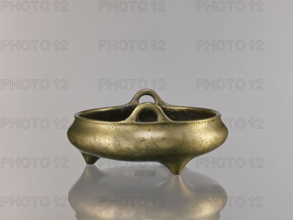 Li-type incense burner, Chinese, bronze ritual vessel, Ming dynasty Xuande period, 1426–1435, China; incense burner; height 10.0 cm, diameter 23.5 cm, A small metallic vessel with a rounded, bowl-like body and a dull gold surface sits on a reflective surface, showing a faint mirror image beneath; the vessel has a curved rim, two arching loop handles that rise from opposite sides and meet near the center, and it rests on three short tapered feet; the metal shows mottled patina and small dark spots, with visible wear and subtle variations in gold, brown, and greenish tones, set against a smooth pale gray background.