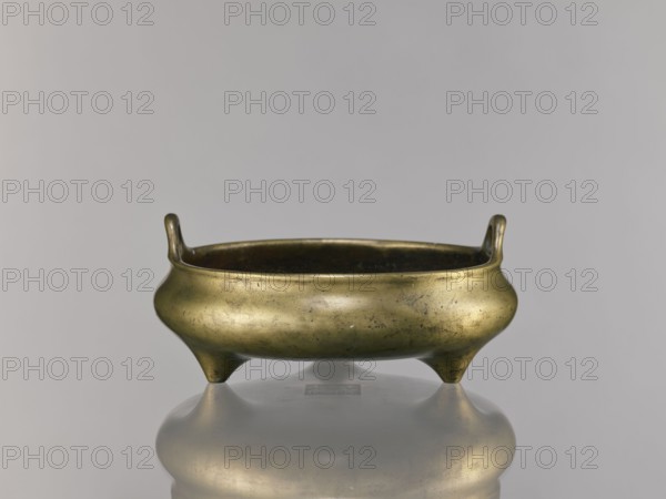 Li-type incense burner, Chinese art; bronze ritual/ceremonial vessel, Ming dynasty, Xuande period 1426–1435, China; height 10.0 cm, diameter 23.5 cm., A low, rounded metal vessel with a golden-bronze color sits on a reflective surface, showing a soft reflection beneath it; the vessel has a wide, shallow bowl shape with a gently flared rim and a pair of short upward-curving handles at opposite sides, and it stands on three short, tapered legs that give it a tripod form; the metal surface shows subtle darker patina spots and worn areas, with variations of golden, bronze, and darker brown tones, and the background is a smooth, neutral gray.