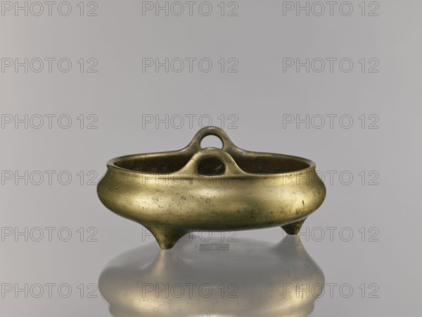 Li-type incense burner, Chinese, bronze ritual/ceremonial vessel, Ming dynasty Xuande, 1426–1435, possibly 1426–1435, China; incense burner; height 10.0 cm; diameter 23.5 cm., A metallic vessel with a rounded, bulbous body and a warm golden-bronze color sits on three short conical feet, showing a few dark spots and subtle surface patina; the rim curves inward and features an arched handle rising from the center with a rounded opening, and the interior appears darker with a greenish-brown tint; the vessel and its reflection rest against a smooth, light gray background with soft shadows.