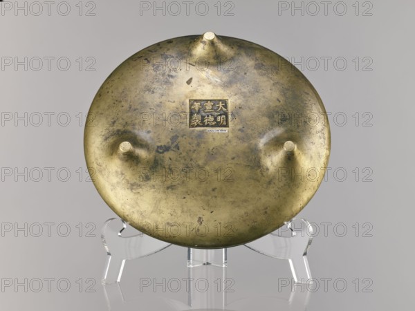 Li-type incense burner, Chinese bronze ritual/ceremonial vessel, Ming dynasty, Xuande period 1426–1435, China; incense burner; height 10.0 cm, diameter 23.5 cm., A round metallic object sits on a clear plastic stand against a neutral gray background; the object has a worn, mottled brass or bronze surface with areas of darker tarnish and lighter polished spots, and it features three raised conical protrusions on one face and a small square recessed area near the center. The plastic stand is transparent and glossy, and the overall color palette includes brassy gold, dark brownish-green tarnish, clear/translucent plastic, and light gray in the background.