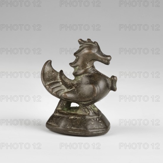 Hintha hintha duck-shaped weight, 10 tikals ticals; Burmese, c. 1760. Bronze weight on an octagonal base marked with a nine-pointed star on the right side. Duck holds a broken twig in its beak. Weight: 317.7 g; height: 6.3 cm., A small sculptural object resembling a stylized animal with a curved tail and raised head sits on a rounded base; the surface shows a patina with shades of brown and green, and the form includes textured ridges and smooth rounded areas, with a hollow opening between the body and the base and subtle irregularities in the metal finish.