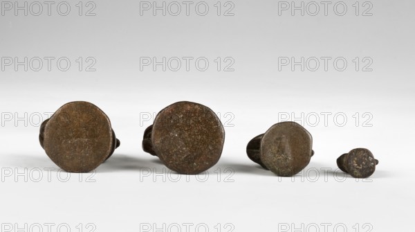 Karaweik weight, 5 tikals; maker, Southeast Asian, Burmese; bird-shaped Karaweik weight on a hexagonal base marked with a drawing of a bird on the right; 2nd half of the 19th century; Burma Myanmar; height 4.35 cm; weight 75.9 g., A row of four round-topped, irregularly shaped metal objects resting on a flat surface, with a smooth light gray to white background that fades to a slightly darker gray toward the top; the objects appear in a gradient of sizes from larger on the left to smaller on the right and show rough, textured surfaces in brown and dark brown with patches of darker greenish-brown patina and small lighter speckles, each casting soft shadows on the surface beneath them.
