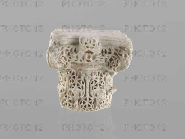 Column capital from Madinat az-Zahra, Islamic/Arabic art, Caliphate of Córdoba 929–1031, Al-Andalus, 10th century; architectural element—capital with acanthus ornament; height 22.3 cm, width 23.3 cm, depth 24.0 cm, weight 15.5 kg., An off-white carved stone fragment with intricate openwork patterns sits against a neutral gray background, showing a weathered top surface and a decorative band with leafy and floral motifs; the carving displays rounded forms, pierced voids, and textured surfaces, and the piece casts a faint reflection on the smooth gray surface below, with visible tones of off-white, light beige, and subtle gray.