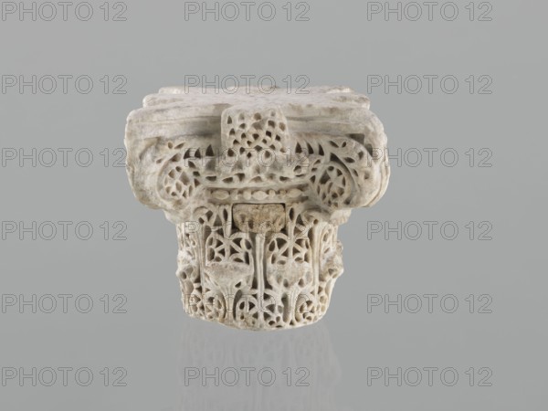 Column capital from Madinat az-Zahra;, Islamic/Arabic art; Caliphate of Córdoba 929–1031, Al-Andalus; architectural element — capital with acanthus ornament; 10th century; place of origin: Madinat az-Zahra, Al-Andalus; dimensions: height 22.3 cm, width 23.3 cm, depth 24.0 cm; weight 15.5 kg., A carved stone architectural fragment sits centered against a smooth grey background, showing intricate openwork and floral-inspired motifs. The piece is predominantly pale beige and off-white with subtle areas of darker beige and light brown discoloration. The top surface is broad and flat with rounded edges, while the lower section narrows and displays a dense pattern of interlacing curves and stylized leaf and petal shapes. The carving creates shadows and small voids that emphasize the three-dimensional detailing, and the stone’s surface appears slightly weathered with fine texture and small pits. A faint reflection of the object is visible on the glossy surface beneath it.