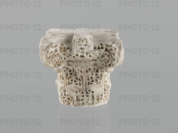 Column capital from Madinat az-Zahra, Islamic/Arabic art, Caliphate of Córdoba 929–1031, Al-Andalus, architectural element capital with acanthus ornament, 10th century, Madinat az-Zahra, dimensions: height 22.3 cm, width 23.3 cm, depth 24.0 cm, weight 15.5 kg., A carved stone object sits against a plain gray background, showing an ornate top and a cylindrical base with openwork floral and foliate patterns. The stone has a pale beige to off-white color with subtle darker beige and gray veining and slight areas of discoloration. The top section is broader and block-like with rounded edges and carved scroll-like details, while the lower section narrows into a columnar form decorated with stylized leaves and flower motifs connected by delicate pierced arches. The surface appears worn in places with soft edges and a matte, slightly textured finish, and a faint reflection of the object is visible on the smooth surface beneath it.