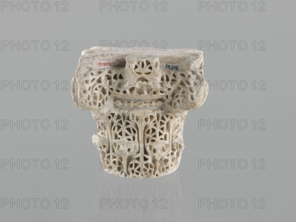 Column capital from Madinat az-Zahra, Islamic art, Arabic art, Arabic culture, Caliphate of Cordoba 929–1031, Al-Andalus, architectural element capital with acanthus ornament, 10th century, Madinat az-Zahra, Al-Andalus; height 22.3 cm, width 23.3 cm, depth 24.0 cm, weight 15.5 kg., A carved stone capital with intricate openwork patterns sits against a plain gray background, showing a weathered surface with varied beige and off-white tones and hints of light brown; the upper blocky section has rounded protrusions and floral-like perforations, while the lower cylindrical portion features vertical, vine-like carvings and leaf-shaped cutouts creating a lace-like appearance; subtle shadows and a faint reflection appear beneath the object, and overall the colors visible are gray, beige, off-white, light brown, and a trace of darker gray.