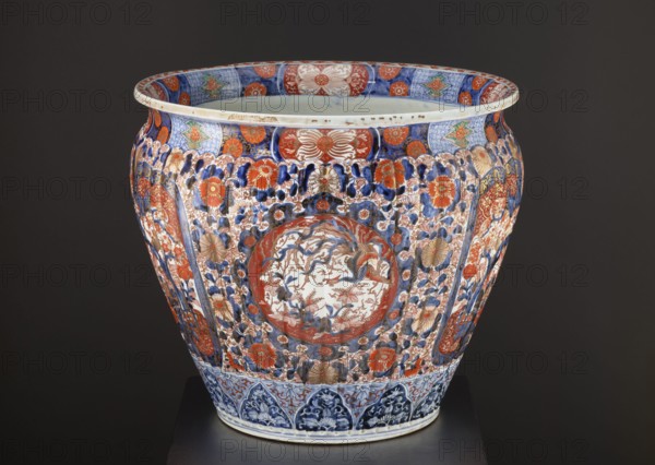 Flowerpot, ca. 1870–1880, Japan Arita. Porcelain jardiniere vessel with colored enamel and Imari palette; height 50.5 cm, diameter 58.0 cm, weight 28.0 kg. Decoration: floral ornaments including chrysanthemums, sakura, peonies, plant tendrils and geometric ornaments; kachôga bird-and-flower imagery featuring phoenixes/hoo and spotted pheasants. Style: Meiji 1868–1912; function: plant ornament., A large decorative ceramic vessel sits against a dark background, showing a bulbous shape with a flared rim and detailed painted ornament. The vessel displays a dense overall pattern of floral motifs and stylized foliage in shades of blue, red, and gold, with areas of white showing through the painted designs. A central roundel on the front contains a more delicate scene rendered in muted blue and red tones surrounded by a red border. Bands of patterned decoration encircle the rim and the base, featuring geometric and floral elements in blue and red with touches of green and gold. The surface al...