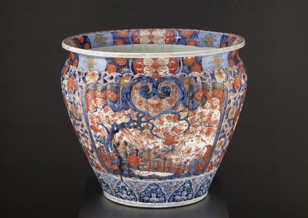 Flowerpot, ca. 1870–1880, Arita, Japan. Ceramic jardiniere colored porcelain, imari, height 50.5 cm, diameter 58.0 cm, weight 28.0 kg. Meiji period 1868–1912. Decoration: floral decorations and plant ornaments—chrysanthemums, sakura, peonies, plant tendrils; geometric ornaments; kachôga and phoenix hoo imagery including spotted pheasants. Function: decorative vessel for plants., A large ceramic vessel with a rounded, slightly flaring rim sits against a dark background; the vessel is richly decorated with intricate floral and scroll motifs in a palette of blue, red, white, and gold, with touches of green; the upper rim interior shows alternating patterned panels in blue and red surrounding a white interior; the body displays ornate cartouche-like panels filled with dense floral scenes and stylized foliage in red and gold on a white ground framed by deep blue scrolling forms; the shoulders and sides feature repeated floral rosettes and decorative borders in blue, red, and gold, and the l...