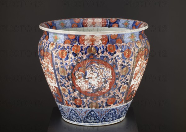 Flowerpot, Japanese, Meiji 1868–1912, ca. 1870–1880. Porcelain jardiniere vessel from Arita, Japan; colored porcelain imari with floral decorations—chrysanthemums, sakura, peonies, plant tendrils—and geometric ornaments. Iconography includes phoenixes hoo, kachôga, and spotted pheasants. Height 50.5 cm, diameter 58.0 cm, weight 28.0 kg., A large ornate ceramic vessel with a rounded shape sits against a dark background, decorated with an intricate pattern of floral and organic motifs in blue, red, orange, gold, and white. The rim interior shows alternating panels of blue and red with floral elements, and the main body is covered in a dense arrangement of stylized flowers and leaves in blue and red with gold accents on a white ground. Central circular medallions feature a more detailed scene in red and blue surrounded by decorative borders, and vertical panels with floral motifs appear on the sides. The lower section has a band of blue-toned decorative arches, and the overall surface sho...