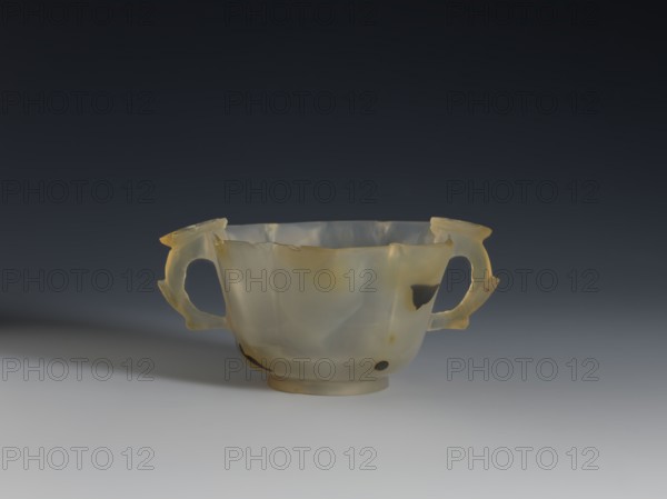 Bowl, 19th century, China; Chinese, Qing 1644–1911. Spotted agate bowl with hexagonal upper part on a ring foot; handles formed as leafy branches with a flower. Morning glory motif. Yellow-rusty stains and black dots in the agate. Vessel; height 4.7 cm, diameter 8.0 cm., A translucent cup with two looped handles sits on a smooth surface against a dark gradient background; the cup is mostly pale cream and milky white with areas of warm yellow and small patches of darker brown, the rim is uneven and slightly jagged, and the handles show thin, delicate contours and subtle variations in opacity, casting a soft shadow on the surface beneath.