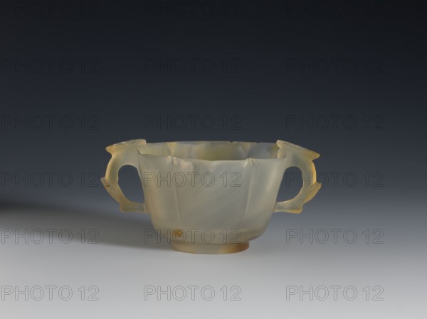 Bowl, 19th century, China, Qing 1644–1911. Spotted agate bowl with yellow-rusty stains and black dots; hexagonal in the upper part, on a ring foot. Handles shaped as leafy branches with a flower. Morning glory motif. Vessel; height 4.7 cm, diameter 8.0 cm., A translucent pale yellow cup with a smooth, slightly marbled surface sits centered on a neutral gradient background; it has a rounded body with a small circular base and two curved handles on opposite sides that are the same translucent pale yellow with slightly darker amber edges, and the rim shows irregularities and subtle chips.