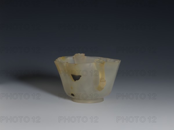 Bowl, 19th century, China, Qing 1644–1911. Spotted agate vessel, hexagonal in the upper part, on a ring foot; height 4.7 cm, diameter 8.0 cm. Decoration: morning glory; handles shaped as leafy branches with a flower. Yellow-rusty staining and black dots in the agate., A translucent cup-like object sits on a flat surface against a dark background, with a smooth rounded base and a slightly flared rim that is irregular and uneven; the object appears made of a milky, semi-opaque material showing creamy white and pale yellow tones with some darker amber areas and a few small darker inclusions; there is a curled handle-like protrusion on one side with a similar pale yellow coloration, and the surface shows subtle variations in translucency and smoothness, casting a soft shadow to one side on the surface below.