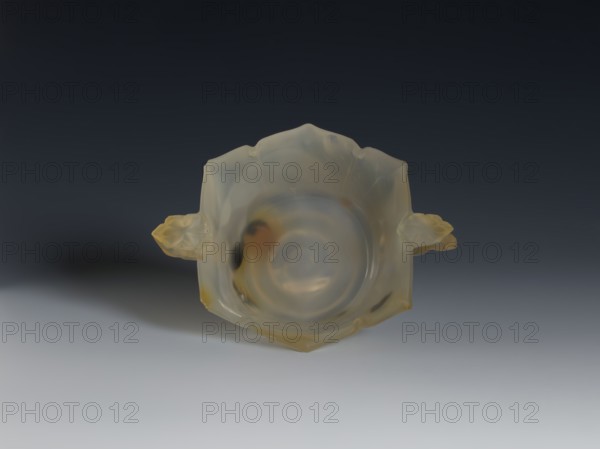 Bowl, 19th century, Qing 1644–1911, China. Spotted agate vessel with yellow-rusty stains and black dots. Hexagonal in the upper part, on a ring foot. Handles shaped as leafy branches with a flower. Morning glory motif. Height 4.7 cm, diameter 8.0 cm., A translucent, pale yellowish bowl with a smooth rounded interior and irregular petal-like edges sits centered against a dark gray to light gray gradient background; the bowl has a glossy surface with subtle milky swirls and warmer amber and honey-colored patches, and a small floral-shaped handle on each side that share the same translucent yellow and amber tones, casting a soft shadow on the lighter foreground.