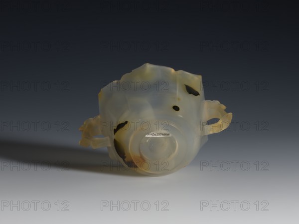Bowl, 19th century, China, Qing dynasty 1644–1911. Spotted agate bowl with yellow-rusty stains and black dots, morning glory motif, hexagonal in the upper part, on a ring foot. Handles shaped as leafy branches with a flower. Vessel; height 4.7 cm, diameter 8.0 cm., A translucent object with a smooth, rounded base and irregular upper edges rests on a flat surface, showing pale milky white and light gray tones with patches of amber and dark brown. The object has a pair of looped handles protruding from opposite sides, also showing the same pale and amber coloration with darker spots. The surface appears slightly glossy, with faint internal striations and subtle variations between milky white, gray, amber, and dark brown areas, and a soft shadow stretches to the left on a gradient background that shifts from light gray at the bottom to dark gray at the top.
