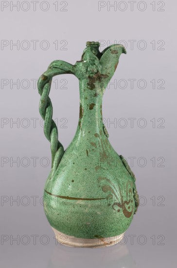 Jug, Islamic Turkish Ottoman dynasty, 1299–1922, glazed ceramic jug with floral ornament, braided handle, filter, curved spout, neck cover with a rosette; 19th century, Çanakkale, Turkey; height 34 cm, diameter 16.2 cm., A green-glazed ceramic ewer with a rounded body, a narrow neck that flares into a pointed spout, and a twisted handle attached from near the spout down to the shoulder, showing wear and spots of brownish discoloration; the surface has darker green speckling and decorative raised motifs on the body and neck, with areas of exposed or lighter ceramic near the base which sits on an unfinished ring.