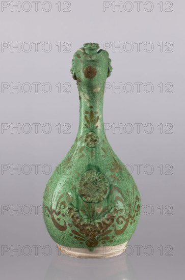 Jug, gift, Islamic art, Turkish culture, Ottoman dynasty 1299–1922, glazed ceramic jug with floral ornament, braided handle, filter, curved spout, neck cover with a rosette, 19th century, Çanakkale, Turkey; vessel, height 34 cm, diameter 16.2 cm., A green ceramic vessel with a long narrow neck and a rounded body sits against a plain light gray background; the vessel has a textured green glaze with darker green and brown decorative motifs including floral and scroll-like patterns, raised circular medallions on the neck and body, and areas of brownish patina or wear; the rim and base show a lighter, unglazed beige edge, and the overall shape tapers from the bulbous lower body to the slender upper neck with a small flared mouth.