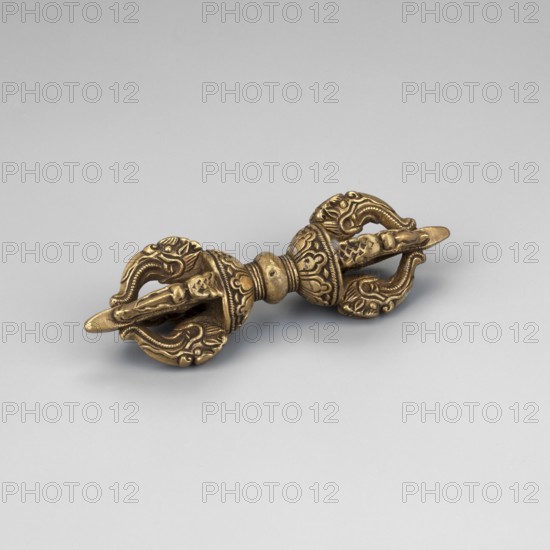 Vajra, Himalayan/Tibetan Buddhist art with Chinese influence; on a lotus base on both sides. Four prongs shaped as dragon heads with flamingly curved tongues, the ends resting on a four-cornered club. In the center of the narrowing is a sphere. 18th–first half of the 20th century; China, / Tibet, ceremonial vajra, height 9.9 cm, width 2.8 cm., A metallic object with a golden-bronze color rests on a smooth light gray surface; it has a symmetrical, double-ended shape with ornate, curved elements and carved floral and wave-like patterns, a central rounded section with a slightly darker patina, and each end features hooked, claw-like protrusions and layered decorative ridges.