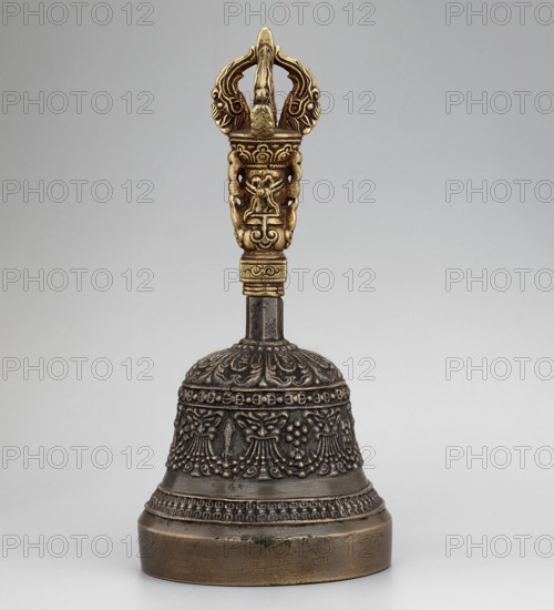 Bell, Tibetan Buddhist art, Himalayan art, Himalayan culture, vajras, bodhisattva iconography, heads, musical instruments, percussion instruments, idiophones, religious rites, ritual object. Bowl with a lambrequin border with mascarons. Handle with a bust of a Bodhisattva carrying a vajra. 18th–1st half of the 20th century, Tibet . Ceremonial object; height 14.9 cm, width 7.15 cm., A decorative metal bell with a dark bronze body and an ornate golden handle stands on a light gray surface against a pale gray background; the bell body shows intricate raised floral and garland patterns and textured bands, while the handle features sculpted, symmetrical motifs and curving shapes with a brighter gold color contrasting the darker bronze stem and base.