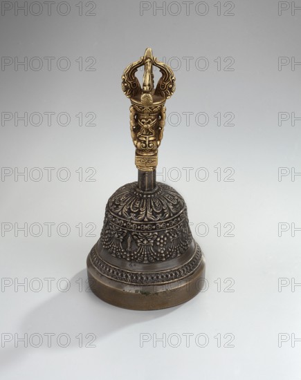 Bell;, Bowl with a lambrequin border with mascarons. Handle with a bust of a Bodhisattva carrying a vajra. Ceremonial object bell. Tibetan Buddhist / Himalayan art; vajras, bodhisattva iconography; heads; musical instruments — percussion idiophone. 18th–1st half of the 20th century; Tibet possibly. Height 14.9 cm; width 7.15 cm., A metal handbell stands on a pale gray background, with a dark bronze bell body decorated with ornate raised floral and patterned designs and a rounded base showing a lighter brownish-bronze rim; the handle is a contrasting golden color with sculpted intricate shapes and curved elements, and the bell casts a soft gray shadow to one side.