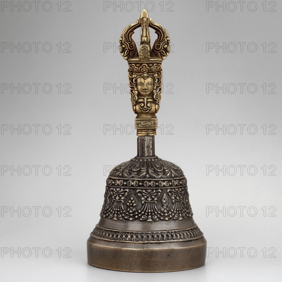 Bowl with a lambrequin border with mascarons. Handle with a bust of a Bodhisattva carrying a vajra. Ceremonial bell. Tibetan Buddhist/Himalayan art; vajras; bodhisattva iconography; heads; musical/percussion instrument idiophone; used in religious rites. 18th–1st half of the 20th century; Tibet place of origin ?. Height 14.9 cm; width 7.15 cm., A decorative metal bell with a dark bronze body and a golden handle is shown against a neutral gray background; the bell body features ornate raised floral and scalloped patterns in dark bronze tones with lighter highlights, a rounded base with a slightly darker rim, and a narrow neck leading to a vertical shaft that connects to an intricately carved golden handle topped by a decorative crown-like element and a stylized human face motif, with visible textures of metalwork and subtle variations of brown, bronze, gold, and gray.
