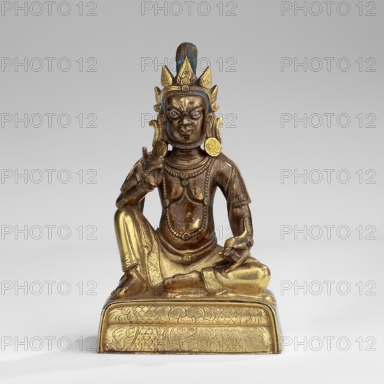 Jambhala, deities of wealth iconography, Himalayan culture, Tibetan Buddhist art, Himalayan art, fruits, mongooses, Yellow Jambhala Tib. dzam b+hA lha — deity of wealth. Shown seated, holding a bijapuraka citrus fruit in his right hand and a mongoose in his left. 18th century, Tibet; sculpture; height 7.7 cm, width 4.7 cm, depth 3.2 cm., A metal statue of a seated figure with a crown-like headdress featuring pointed elements and a tall central plume, rendered in brown and gold tones with touches of blue and green; the figure wears ornate jewelry and garments, has a detailed face with pronounced features, and holds one hand raised while the other rests near the knee; the figure is seated on a decorated rectangular base with engraved patterns in gold, set against a plain light gray background.