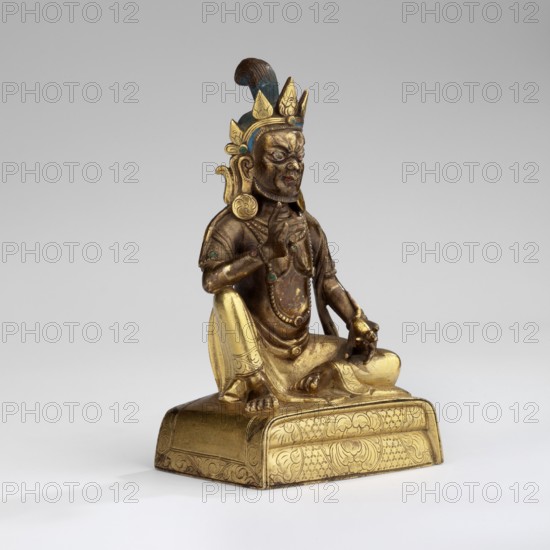 Jambhala, unknown sculptor, deity of wealth iconography, Himalayan/Tibetan Buddhist art, fruits, mongooses, Yellow Jambhala Tib. dzam b+hA lha — deity of wealth. Shown seated, holding a bijapuraka citrus fruit in his right hand and a mongoose in his left. 18th century, Tibet; sculpture. Height 7.7 cm, width 4.7 cm, depth 3.2 cm., A seated metal figure with a golden and brown surface wearing a crown with blue and gold elements sits on an ornate rectangular base that is gold in color; the figure has visible jewelry and beads with small blue-green accents, a blue feather or plume behind the crown, and detailed facial features and carved hair that appear darker brown, while the clothing and base show polished gold tones and engraved patterns.