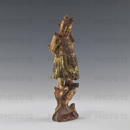 Guardian of Heaven, figurine, art of Southeast Asia, Vietnamese culture, 1st half of the 19th century, Vietnam, sculpture; height 22.8 cm, width 7.2 cm, depth 6.2 cm, A carved standing figure with ornate armor and a headdress, displayed against a neutral gray gradient background; the figure and its pedestal show a worn surface with visible patches of gold and dark brown, along with areas of reddish brown and traces of black, and the carved base has flowing, scroll-like forms continuing the same color palette.