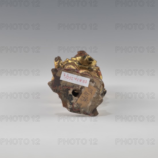 Guardian of Heaven, figurine, art of Southeast Asia, Vietnamese culture, 1st half of the 19th century, Vietnam, sculpture; height 22.8 cm, width 7.2 cm, depth 6.2 cm, The image shows a small sculptural object placed on a flat surface against a smooth gradient gray background. The object has a worn, textured base with a rough, irregular shape and a visible central hole. The visible surfaces display metallic gold areas with dark tarnish, along with patches of reddish-brown and earthy beige tones. There is also a small area of pale off-white on the base and traces of darker blackish spots. The lighting is soft, casting a faint shadow beneath the object.