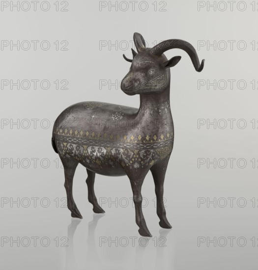 Ram, Persian, Qajar dynasty 1785–1925, sculpture; 1st half of the 19th century, Iran. Material: not specified. Subject: ram; animal figurine with horns. Decoration: decorated on the back with medallions and palmettes with floral ornament; geometric motifs and floral ornament. Dimensions: height 22.5 cm, width 20.5 cm, depth 9.0 cm; weight 0.64 kg., A sculpted animal figure resembling a goat or antelope stands on a smooth reflective surface, shown in profile with its head turned slightly toward the viewer; the body is a dark metallic gray with a weathered patina and decorative etched patterns in muted gold and lighter gray across its torso and neck; the head has curved horns, small upright ears, and subtle engraved facial markings; the legs are slender and slightly stylized, casting faint reflections on the pale neutral background which is an off-white to light gray color.