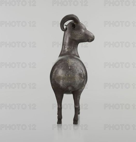 Ram, Persian, Qajar dynasty 1785–1925, 1st half of the 19th century. Sculpture: animal figurine of a ram. Materials/technique: decorated on the back with medallions and palmettes with floral ornament; geometric motifs; horns. Dimensions: height 22.5 cm, width 20.5 cm, depth 9.0 cm; weight 0.64 kg. Categories: Islamic art; sculptures; animals; figurines; rams., A dark metallic sculpture of an animal-like figure standing on a reflective surface, shown from the rear three-quarter view, with a rounded body, slender legs, and a long neck rising to a stylized head with curled horns; the surface shows decorative etched spiral and geometric patterns in lighter metallic tones, with areas of subtle wear and patina; the overall colors visible are dark gray, silver, and hints of muted gold against a pale gray background.