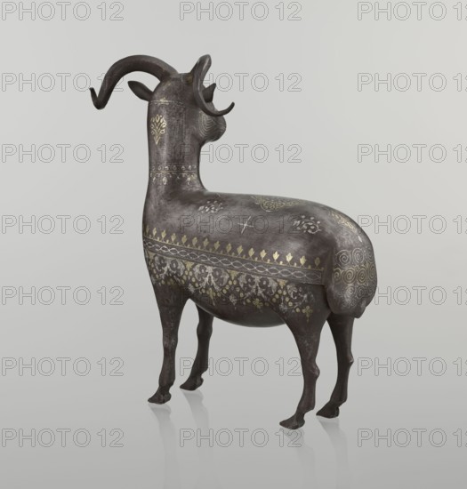 Ram, Persian, Qajar dynasty 1785–1925, 1st half of the 19th century. Sculpture; animal figurine of a ram. Decorated on the back with medallions and palmettes with floral ornament; geometric motifs and floral ornament; horns., Islamic art. Dimensions: height 22.5 cm, width 20.5 cm, depth 9.0 cm; weight 0.64 kg., A sculptural animal figure standing on a reflective surface, shown from a rear three-quarter view with its head turned slightly to the side; the figure has curled horns and slender legs and a rounded body decorated with ornate patterns. The surface of the figure is primarily dark brown with areas of lighter brown patina, and it is embellished with metallic gold and pale silver decorative motifs in floral, geometric, and spiral designs. The background is a smooth gradient of light gray tones.